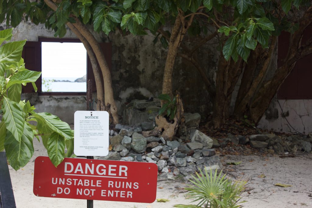 Photograph of overgrown ruins in Virgin Islands National Park. A sign reads "Danger: Unstable Ruins. Do not enter." A rock formation in the ocean is faintly visible through an open window in the ruins.